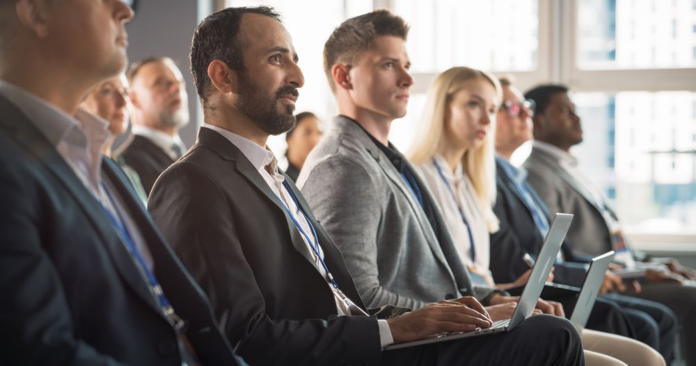 Multiethnic Man Sitting in a Crowded Auditorium at Conference. Middle Eastern Businessman Listening To Keynote, Typing On Laptop. Specialist Watching Technology Presentation About Software Solutions.