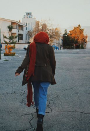 Back view of woman in brown coat and blue denim jeans running on gray asphalt road