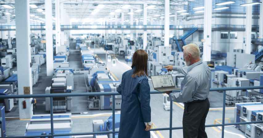 Two Multiethnic Engineers Standing on a Platform, Using Laptop Computer at an Electronics Factory. Machines are Undergoing Maintenance, Specialists Monitoring the Progress Through Online Software