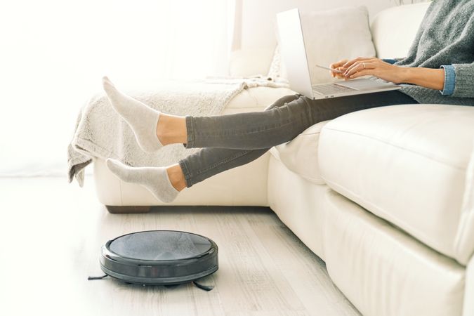 Female freelancer raising legs above working robotic vacuum clea