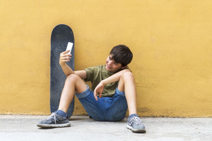 Front view of boy sitting on ground leaning on a yellow wall and taking a selfie