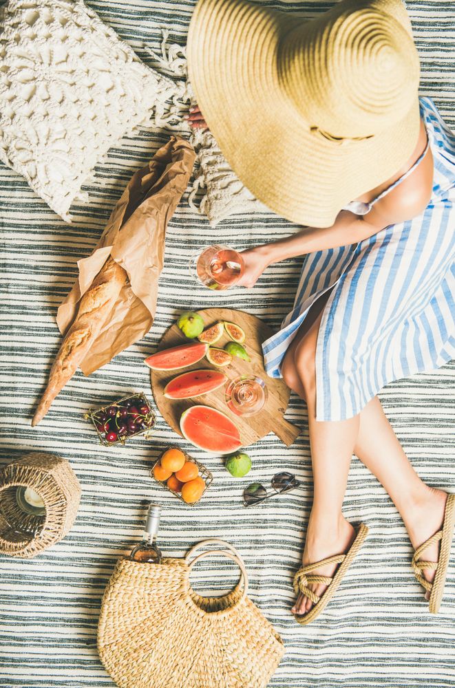 Woman lounging on picnic blanket with large hat thatched bag with glass of rose, baguette, fruit
