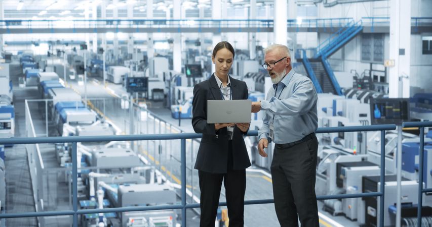 Male Engineer and a Female Technician Having a Conversation and Using Laptop Computer. Specialists Monitoring, Analyzing Conditions at a Modern Electronics Factory with Automated Robotic Arms