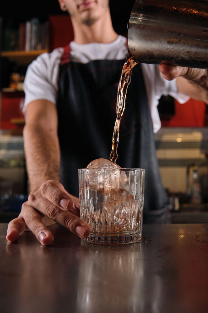Crop barman pouring drink into glass with ice cubes
