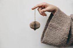 Female hand in woolen sweater holding paper Christmas ornament on plain background