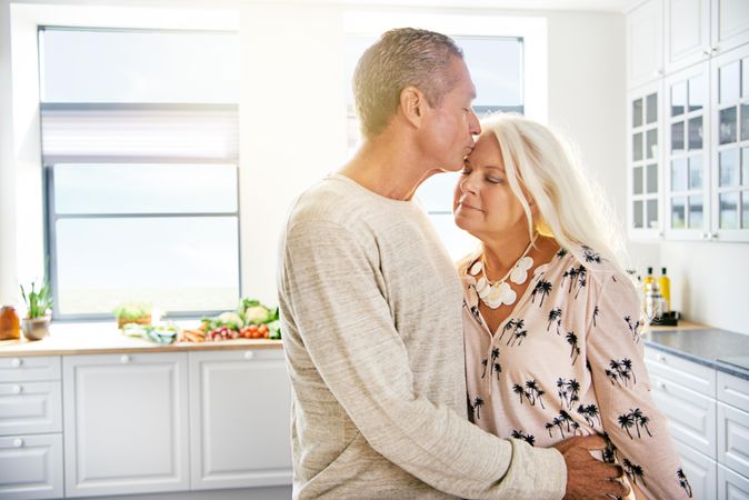 Older man kissing wife’s forehead in bright kitchen