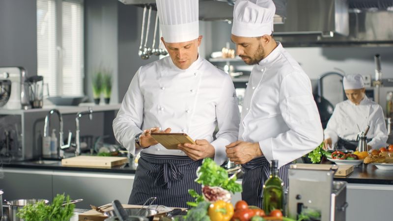 Two Famous Chefs Exchange Ideas About Video Blog Recipe they Watching on Tablet. They are Working in Big Restaurant Kitchen.