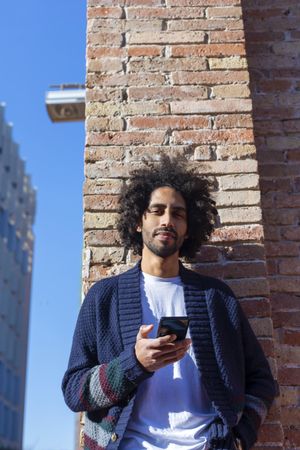 Smiling Black male holding his smartphone while leaning on a brick wall outdoors on sunny day