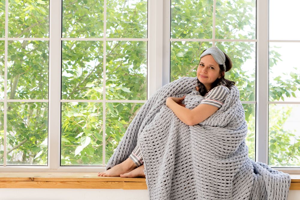 Woman smiles warmly, wrapped in chunky gray blanket by window.