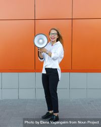 Laughing woman in glasses in front of orange wall with into megaphone 56rWY5