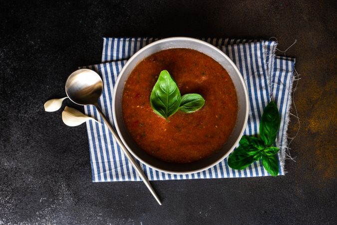 Grey bowl of gazpacho soup with basil leaves on striped napkin