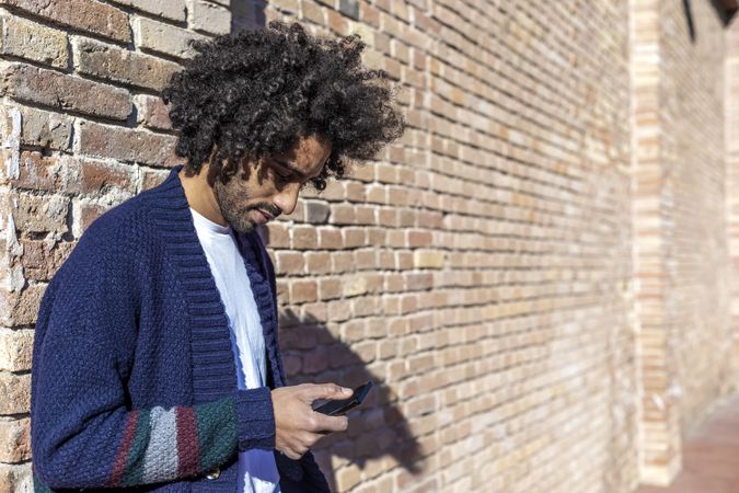 Curly haired male using his smartphone while leaning on a brick wall outdoors on sunny day