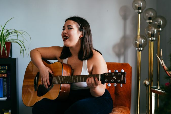 Young woman singing and playing guitar in house sitting on orange chair