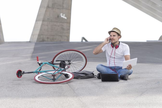 Male sitting outdoors on phone with bicycle and using laptop