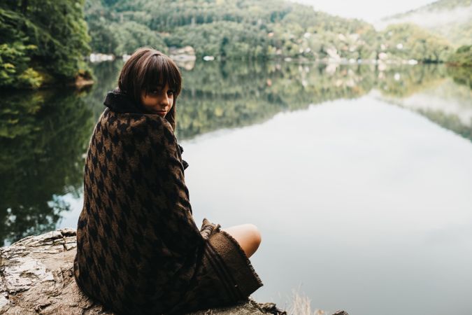 Woman sitting with blanket near lake and mountains