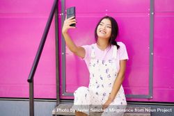 Smiling Chinese woman talking on video call on smart phone  sitting in front of pink trailer 5q7apb