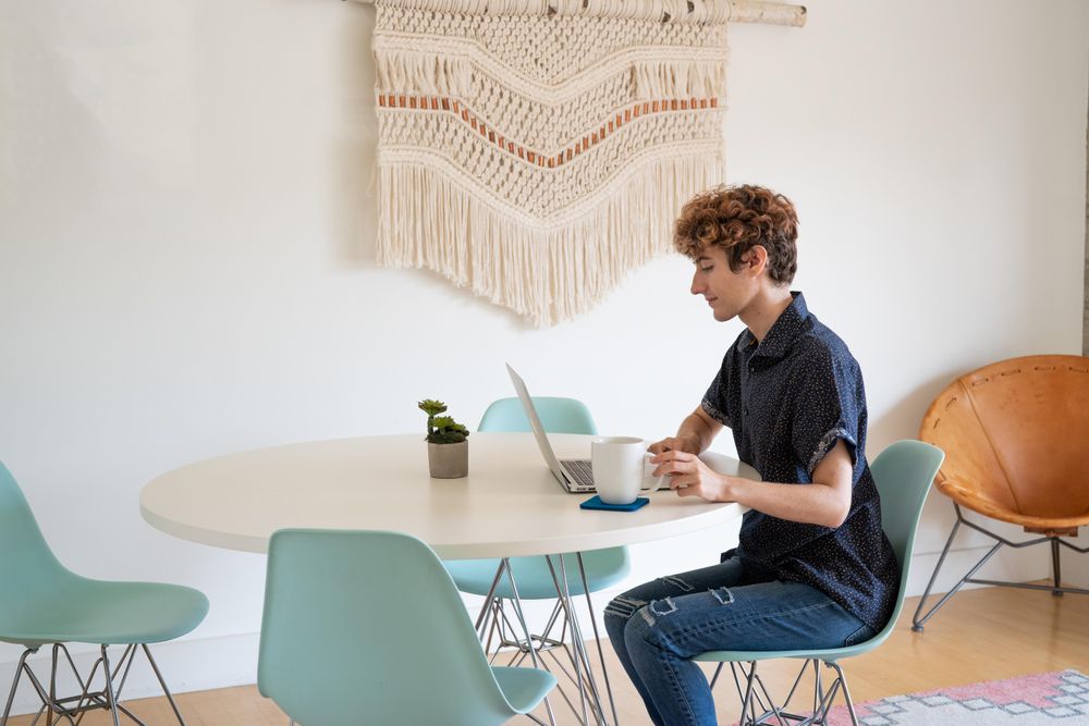 Nonbinary person reaching for coffee while working on a laptop