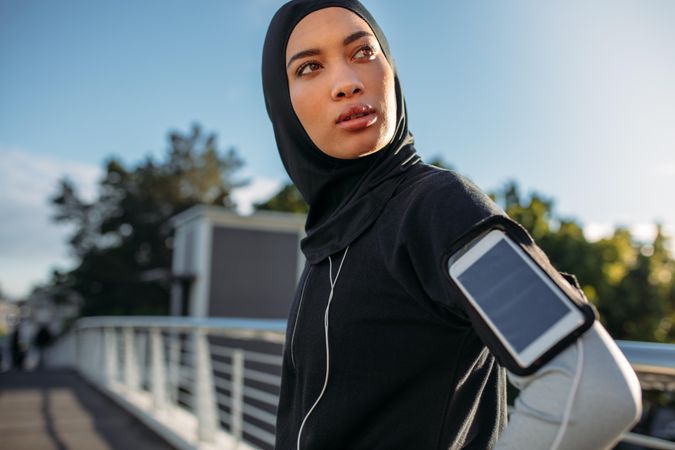 Young sportswoman in hijab standing outdoors with mobile phone on her armband looking away