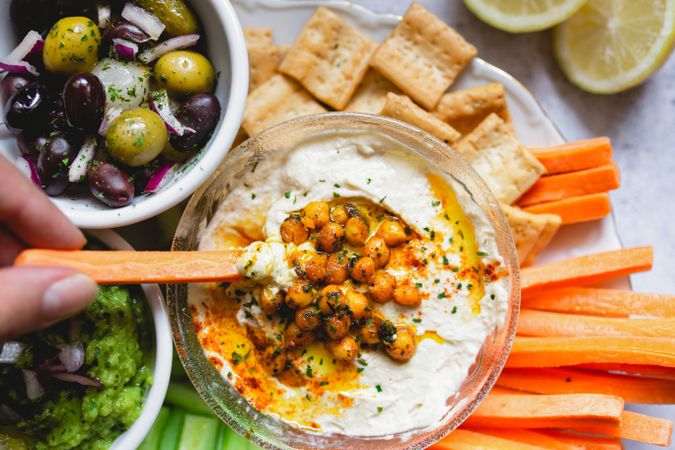 Person dipping carrot stick into hummus decorated with vegetables