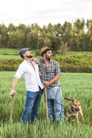 Two men in plaid shirt standing in long grass with forest in background with dog