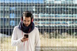 Young man walking in scarf leaning back on metallic fence and looking down at smartphone bGRKrB