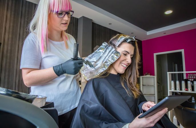 Client smiling looking at digital tablet while having her hair dyed in salon