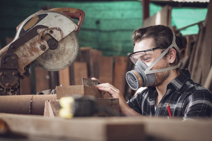 Male carpenter wearing protective mask using electric circular saw cutting wood board