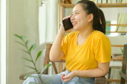 A young woman sits in a chair talking on the phone in the living room at home.