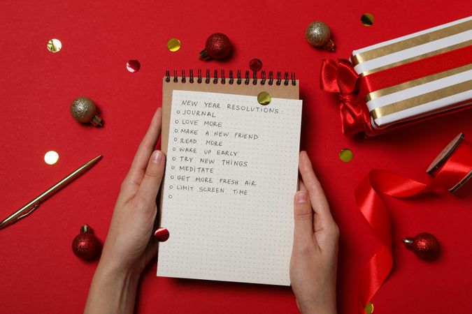 Notepad with list in female hands and decorations on red background, top view