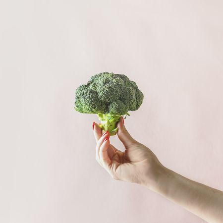 Woman hand holding whole raw green broccoli at pale beige wall background