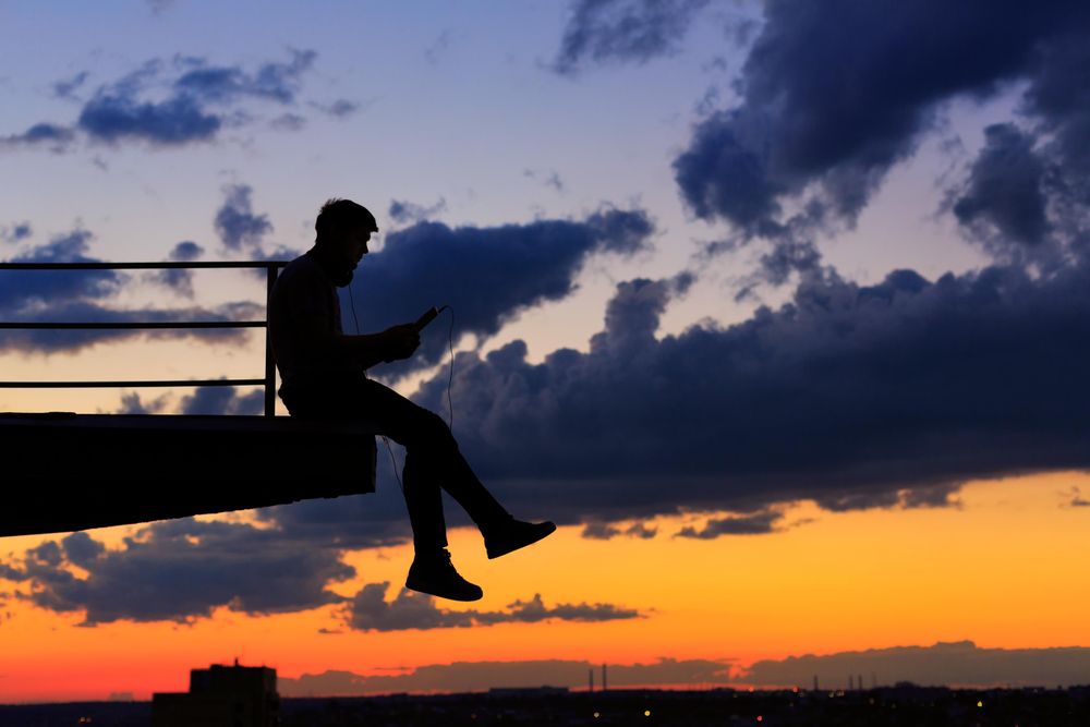 Out line of male sitting on ledge with tablet against a colorful dusk side, vertical