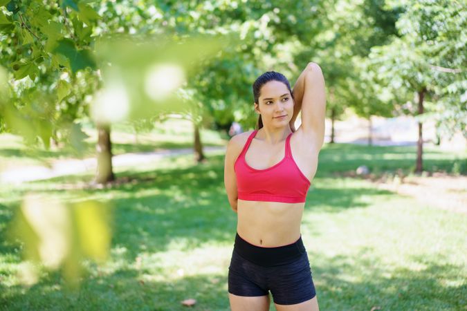 A woman in activewear is stretching outdoors in a park surrounded by greenery, under the warmth of the sunlight