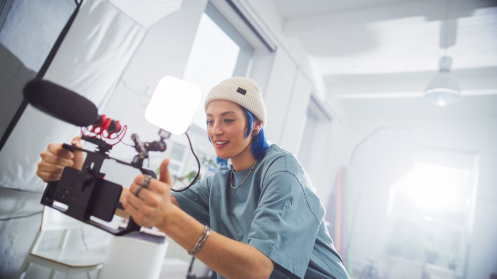 Young Female Filmmaker With Blue Hair, Wearing A Beanie, Operates A Professional Camera In A Well-lit Studio, Capturing Engaging Content For A Creative Advertising Project.