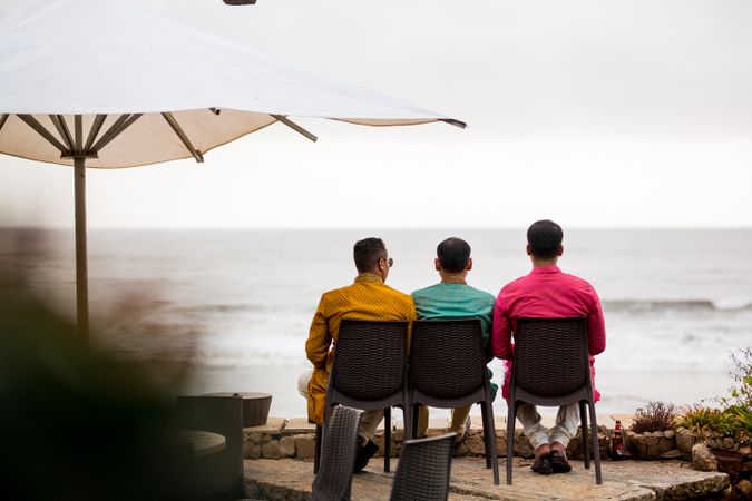 Back view of three men wearing colorful shirts sitting on plastic chairs at the beach