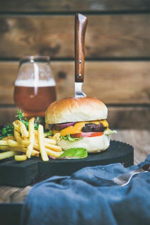 Classic hamburger skewered with knife, with fries and beer at wooden restaurant table