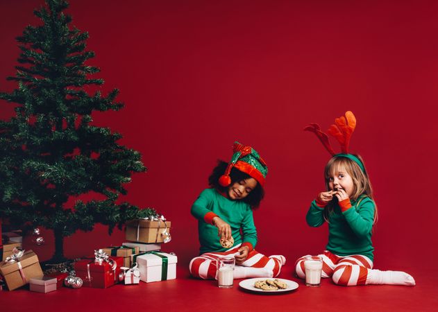 Cute little girls wearing Christmas pajamas and hats eating cookies with milk