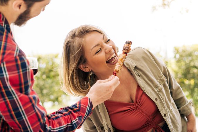Woman laughs as she eats skewers of meat from her friend's hands during a trip to the countryside