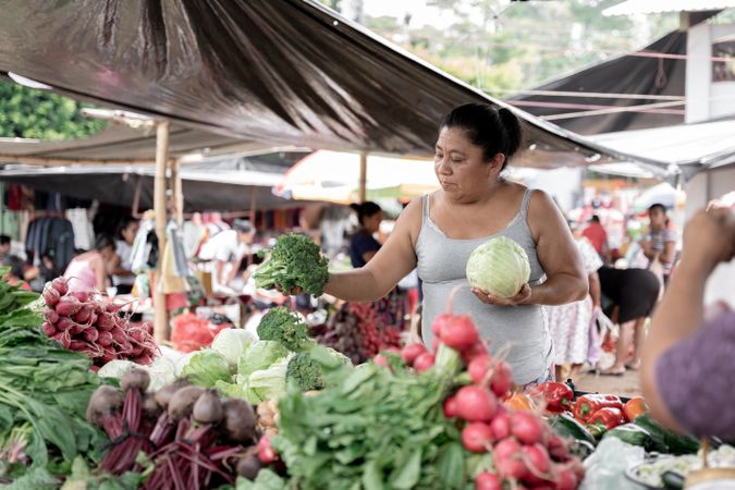 An adult Hispanic woman is holding a cauliflower and a brocoli in a street market