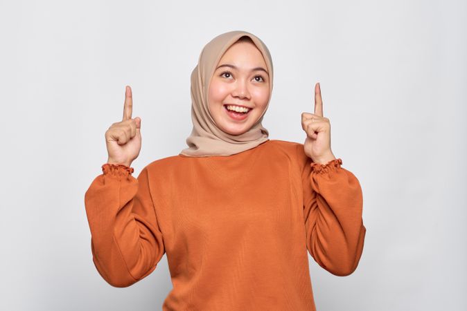 Happy Muslim woman in headscarf and orange sweater pointing upwards with bright idea