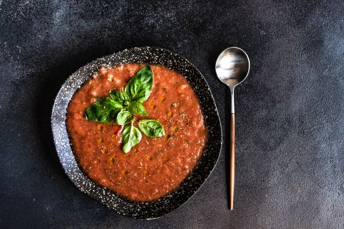 Top view of large bowl & spoon with Spanish gazpacho with basil leaf garnish and salt in dark bowl on counter