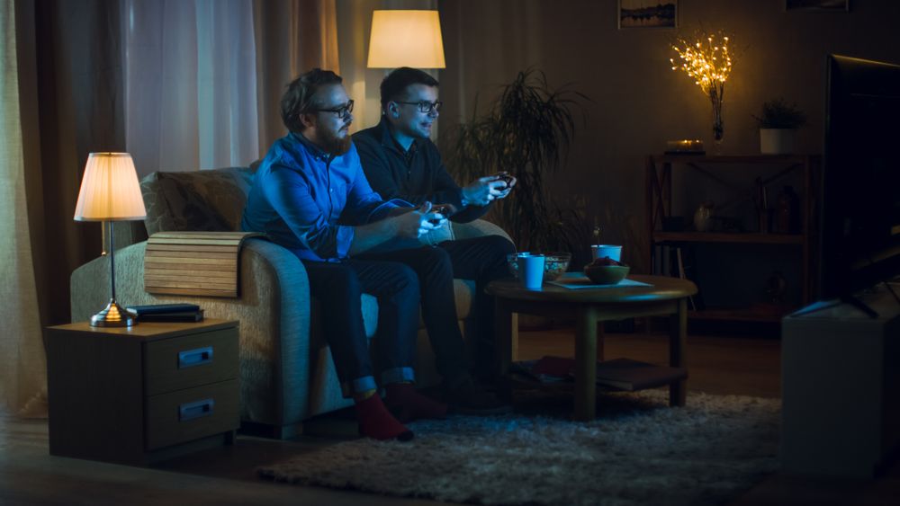 In the Evening Two Friends are Sitting on a Sofa in the Living Room and  Playing Competitive Video Games.