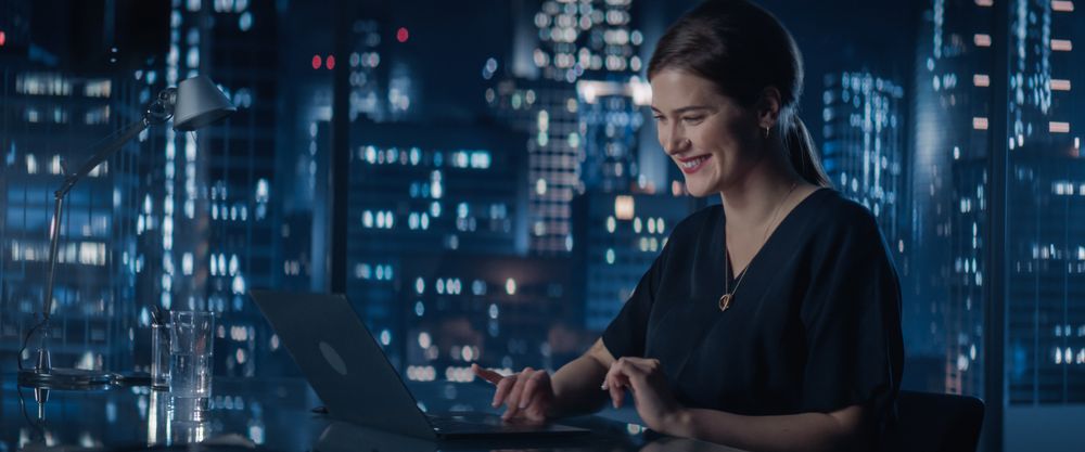 Portrait of Successful Young Businesswoman Sitting at Her Desk Working on Laptop in Big City Office Late At Night.