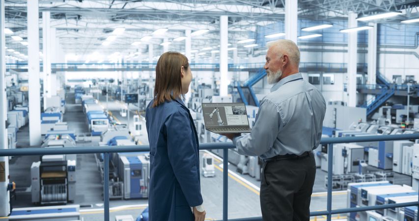 Mature Engineer and Young Japanese Technician Having a Conversation and Using Laptop Computer. Specialists Monitoring, Analyzing Conditions at a Modern Electronics Factory with Automated Robots