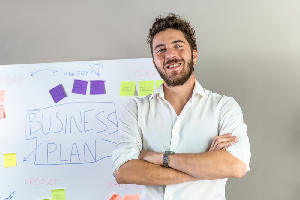 Confident young entrepreneur standing in front of a business plan whiteboard.