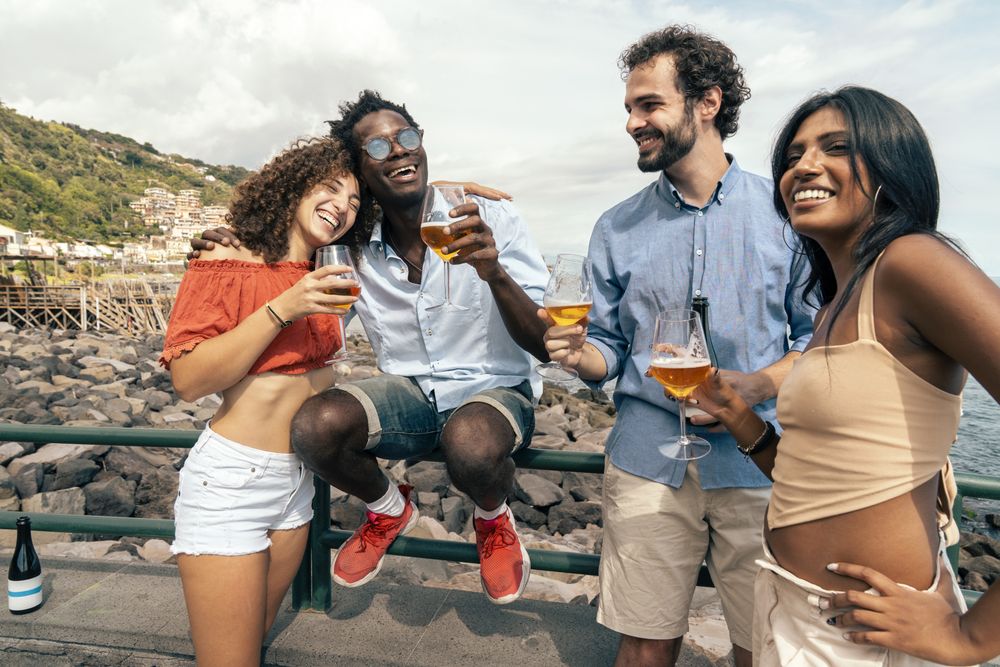 Joyful multicultural friends laughing and embracing by the sea with craft beer.