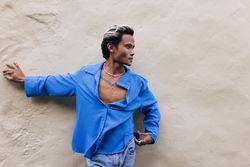 Male in blue shirt and jeans leaning against wall and looking to his side