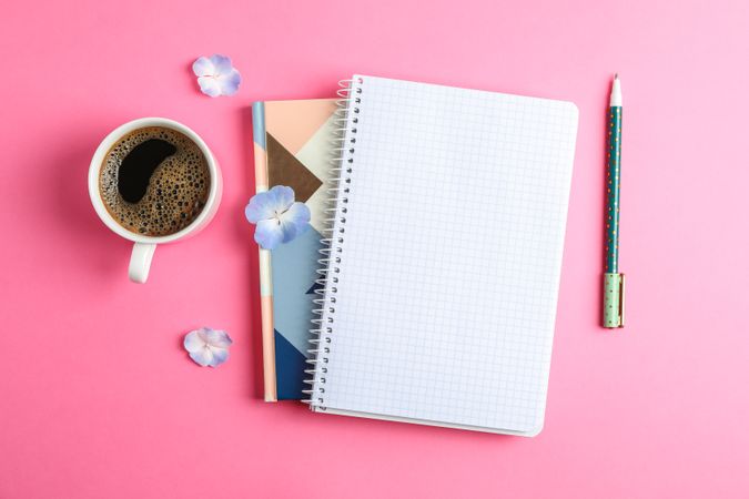 Cup of coffee with petals of flower, copybooks and pen on color background, top view