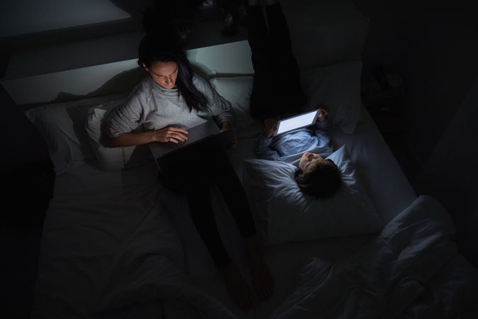 Woman and boy lying on bed and surfing gadgets at night