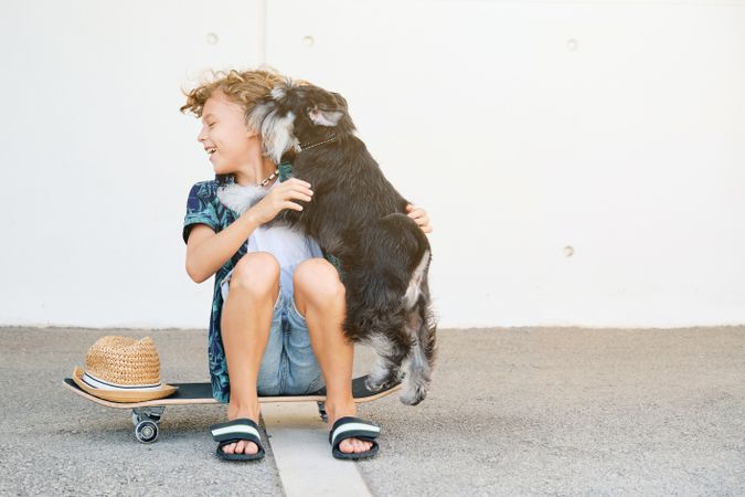 Dog licking face of boy sitting on skateboard