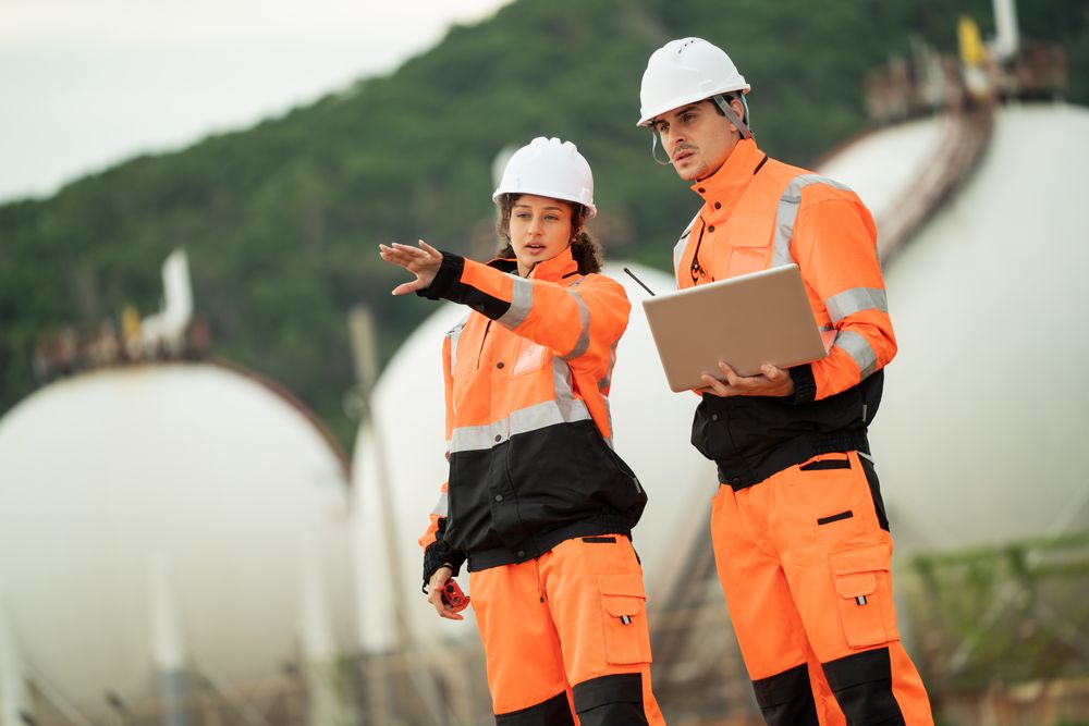 Two engineers in safety gear talking work near spherical gas storage tanks.